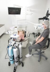 A young girl smiling in a dental clinic with a dentist beside her, showcasing a friendly and professional healthcare setting.